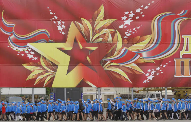 Schoolchildren stand in front of a huge red and yellow flag and coat of arms.