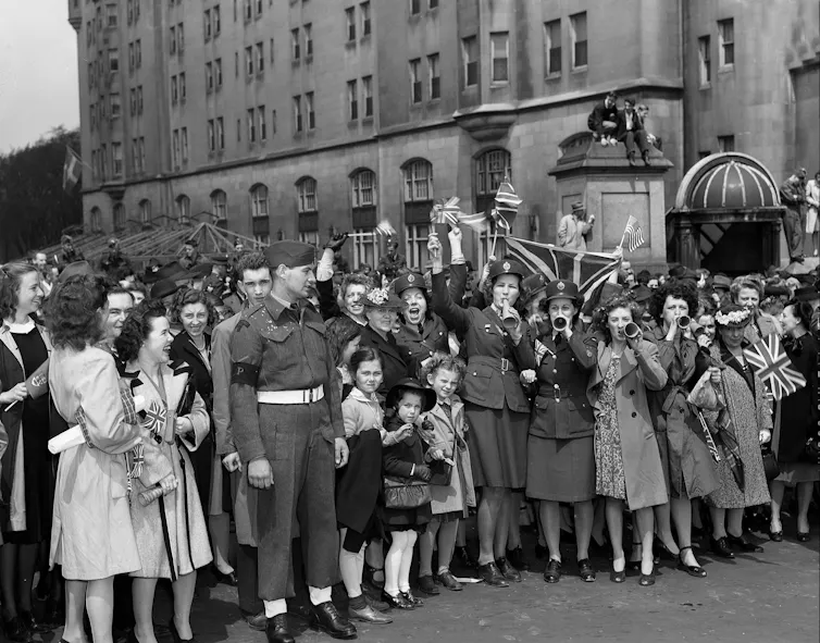 A group of citizens, mostly women and children, smile and cheer in a black and white photo.
