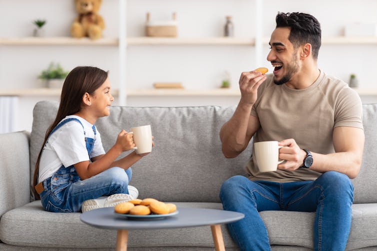 A man and girl seated at different ends of a couch with mugs.