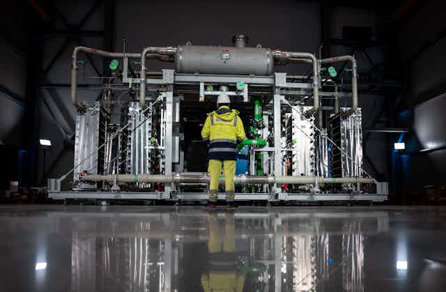 A person in high-vis overalls stands in front of an electrolyser that produces green hydrogen