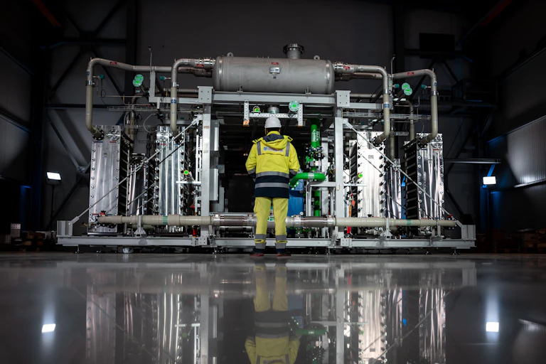 A person in high-vis overalls stands in front of an electrolyser that produces green hydrogen