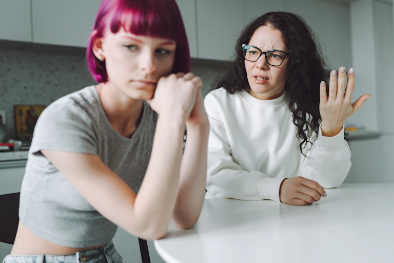 An upset-looking mother and stressed-out teenage daughter sit at a kitchen table. The mother is speaking to the daughter, who is looking away.