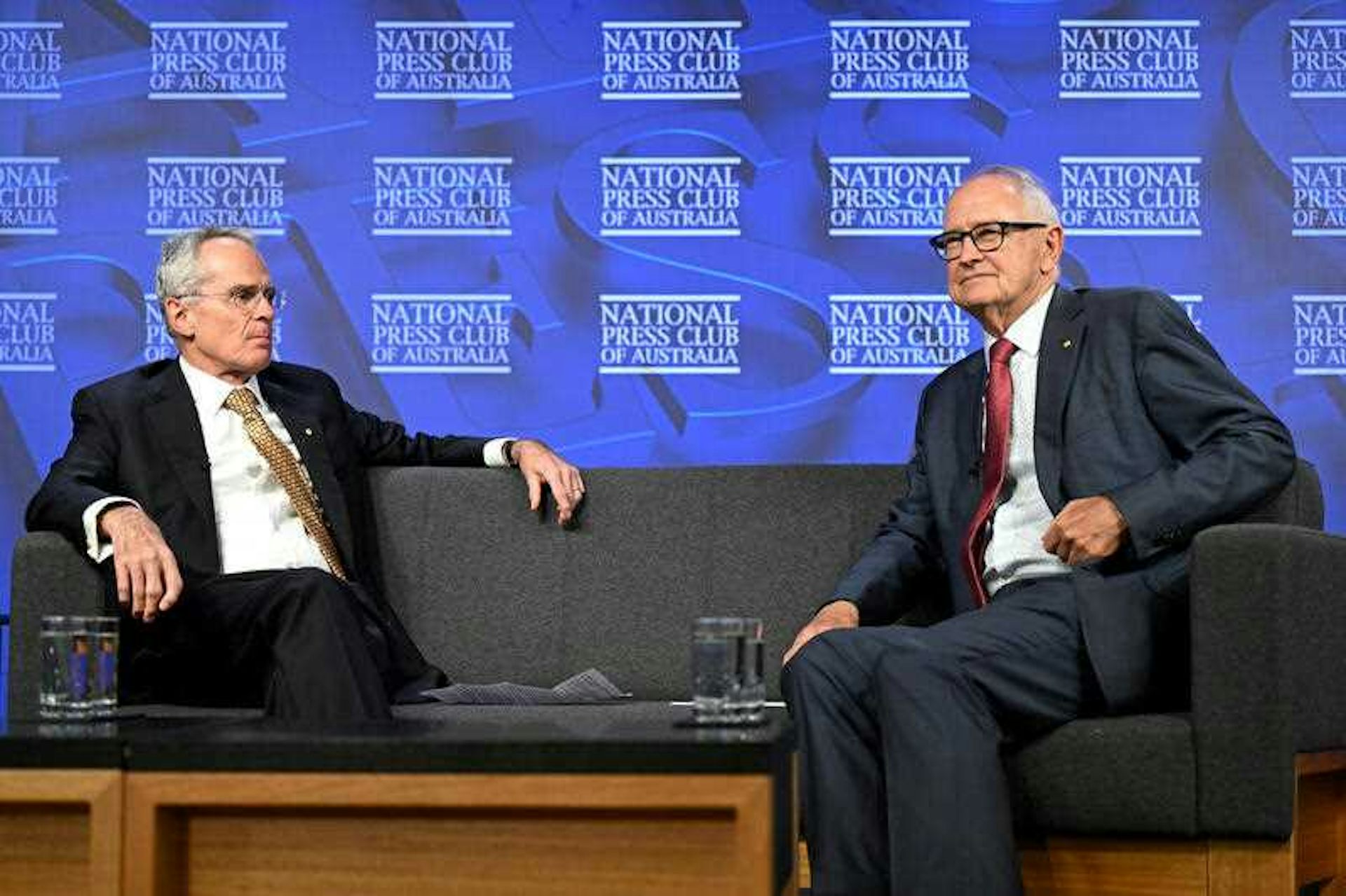 two men sitting on couch in front of blue background