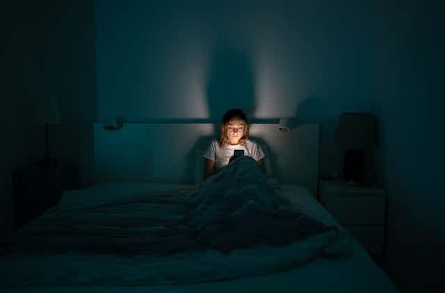 Photo of a woman sitting in bed in a dark room, lit only by the glow of her phone.