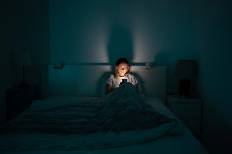 Photo of a woman sitting in bed in a dark room, lit only by the glow of her phone.