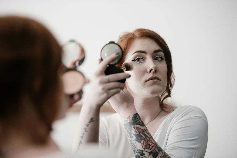 A redhaired woman applies makeup while looking in a compact mirror.
