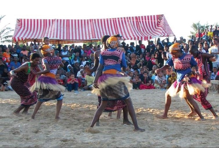 Six women dance in a circle, wearing traditional attire and grass skirts. A crowd watches them from a stand.