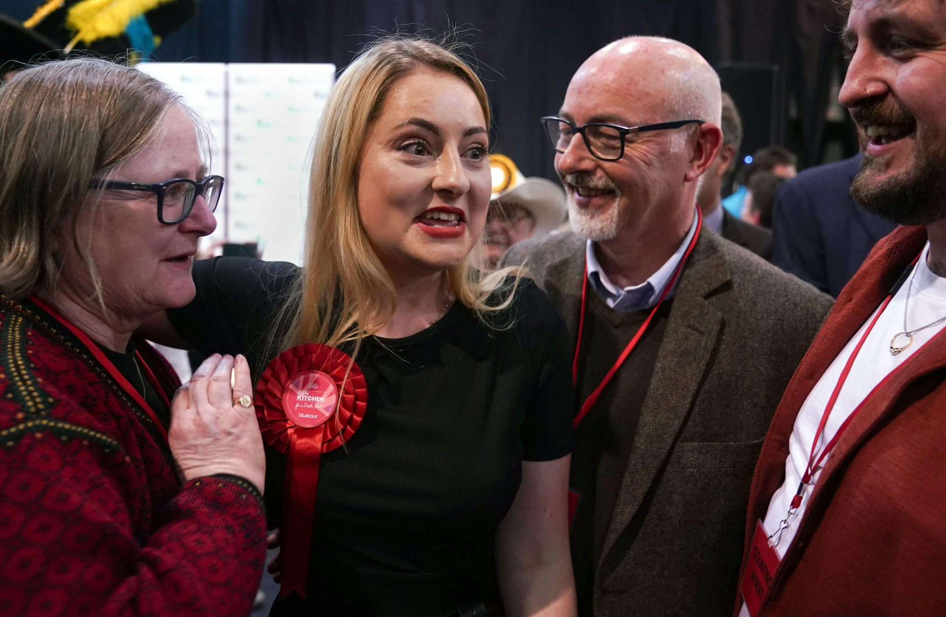 Gen Kitchen, a young, white woman with blonde hair, wearing a Labour rosette, has an expression of triumphant surprise on her face, surrounded by her smiling parents as she is declared winner of the byelection