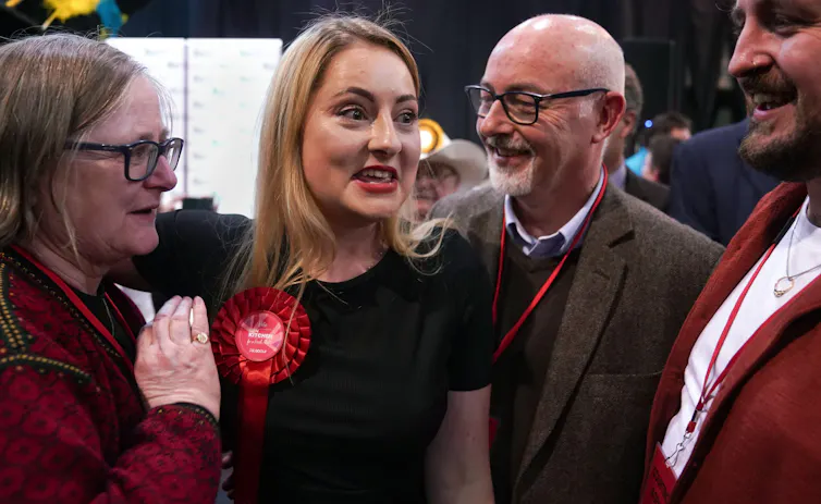 Gen Kitchen, a young, white woman with blonde hair, wearing a Labour rosette, has an expression of triumphant surprise on her face, surrounded by her smiling parents as she is declared winner of the byelection