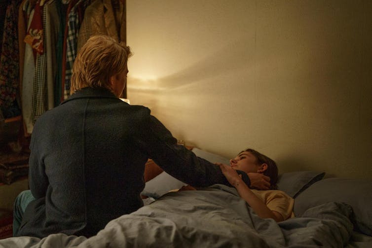 Una mujer está tumbada en la cama casi a oscuras mientras un hombre sentado a su lado le acaricia la cara.