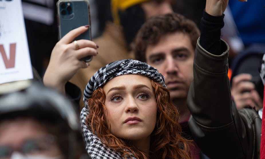 A young woman with long red hair, wearing a black and white scarf wrapped around her head, holds up a mobile phone to record at a protest