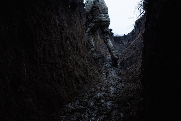 A man in military fatigues peers over the top of a muddy trench.