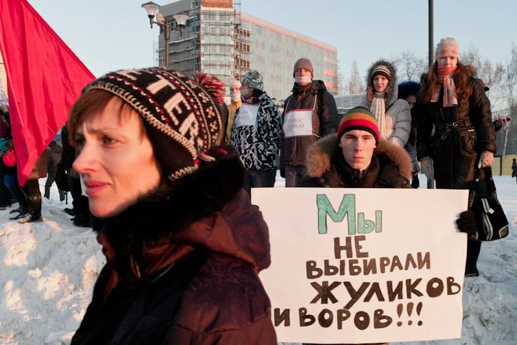 A protester wearing a hat stands in front of a sign in Russian that translates to 'We did not vote for crooks and thieves!'