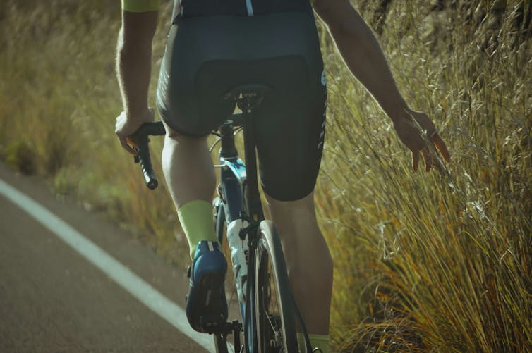 Close-up of lower half of the back of a person cycling, one hand outstretched towards the vegetation on the side of the road