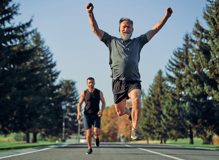 Two people running on a road lined with trees -- the younger person is trailing behind the older person who has leaped into the air with arms raised