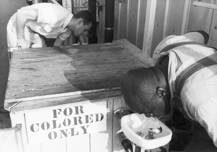 A Black man is drinking from a water fountain that has a signs that reads for colored only.