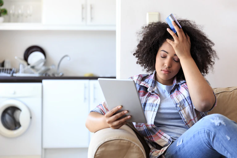 A young woman holds an iPad in one hand and her credit card in between the fingers of another. She's frustrated and she's placed the palm of the hand that has the credit card in her fingers on her head.
