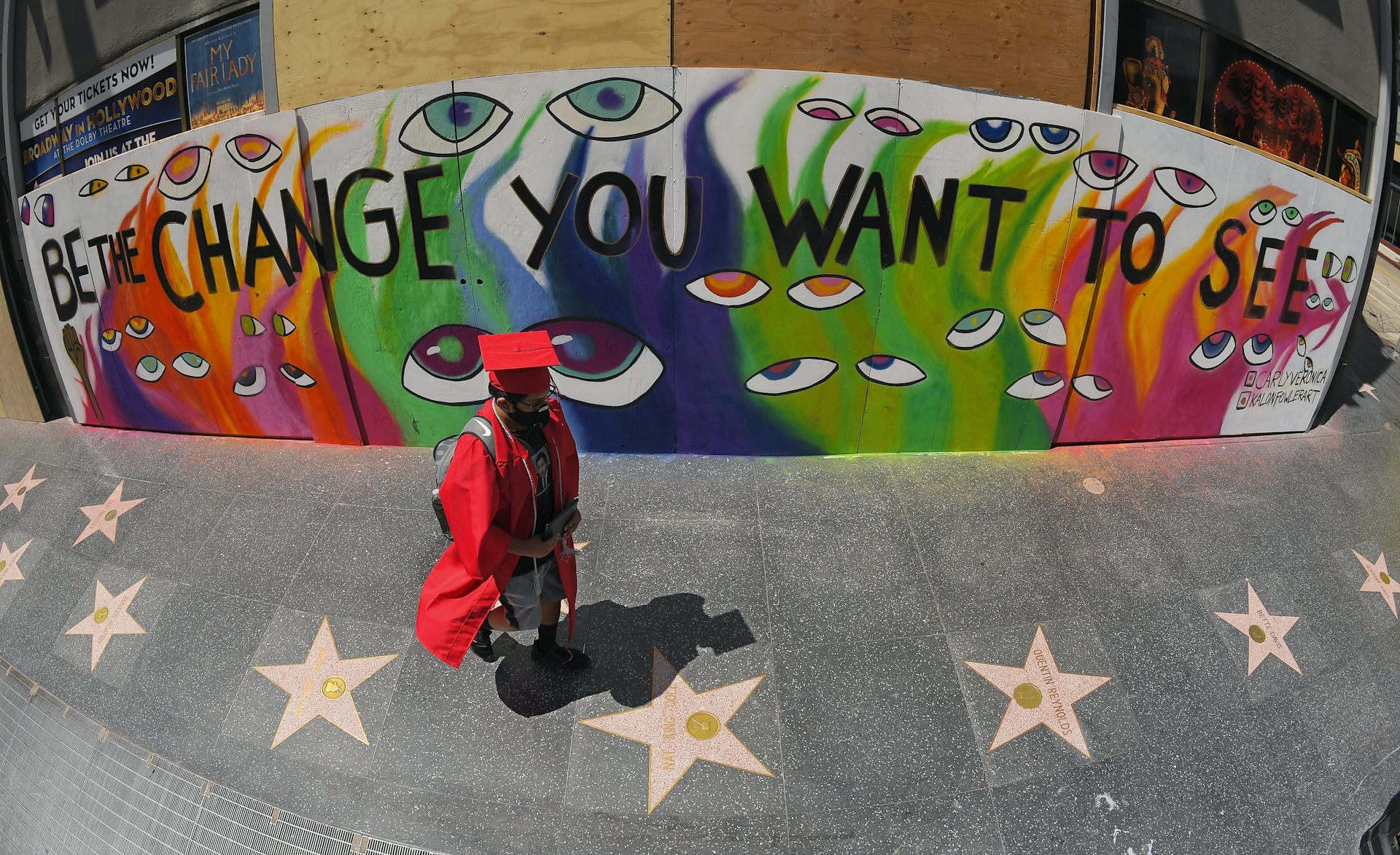 A person in cap and gown in front of mural that says 'be the change you want to see.'