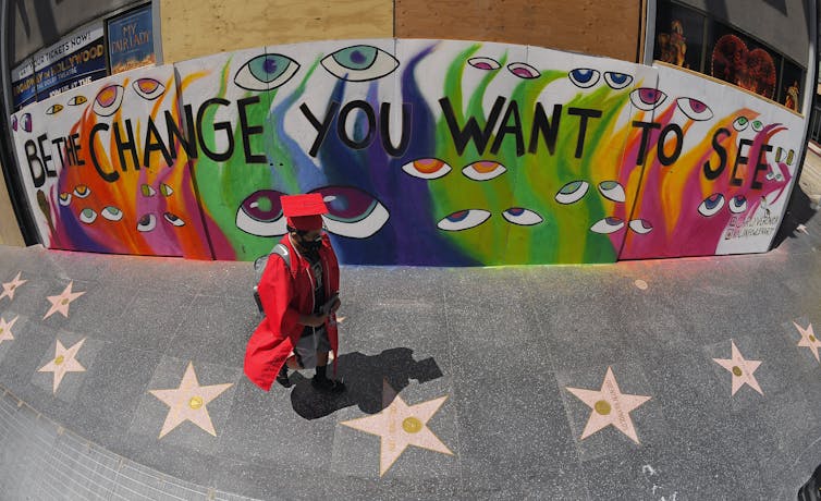 A person in cap and gown in front of mural that says 'be the change you want to see.'
