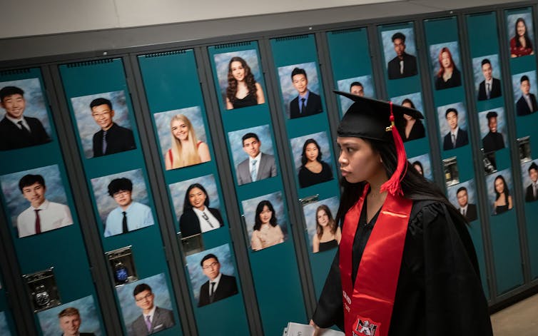 A student seen in graduation cap and gown in front of photos of students on lockers.