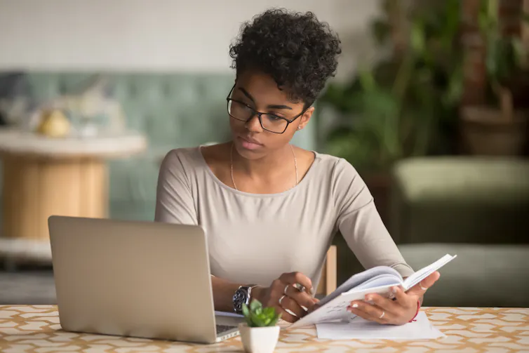 A woman works on a laptop holding a book in her hand.