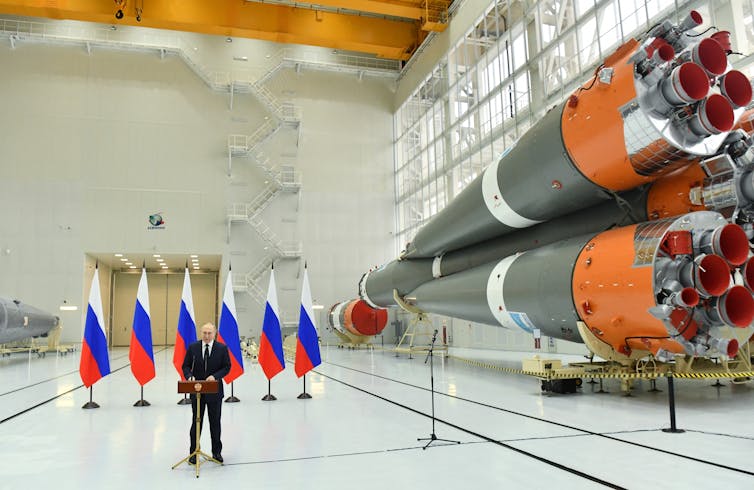 A man stands in front of red, blue and white flags and next to large rockets.