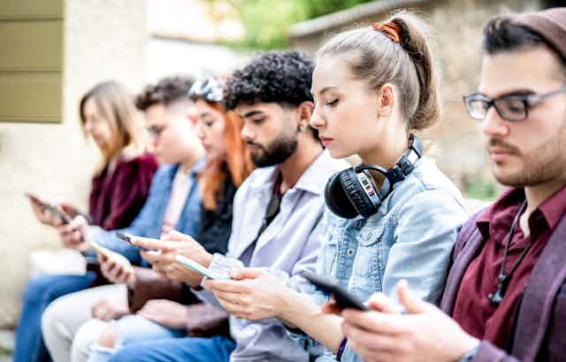 Um grupo de jovens senta-se em um banco enquanto visualiza e envia mensagens de texto em seus smartphones.