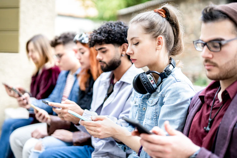 Um grupo de jovens senta-se em um banco enquanto visualiza e envia mensagens de texto em seus smartphones.