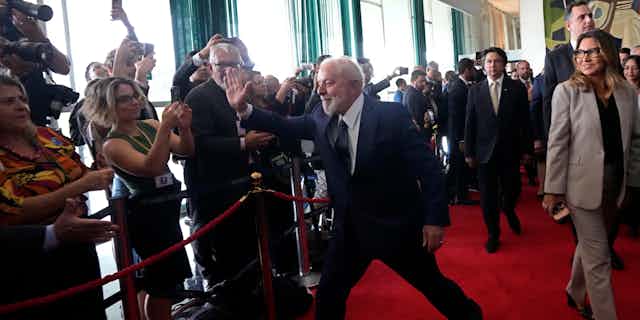 A man with white hair and beard in a suit and tie high fives members of the media while walking down a red carpet.