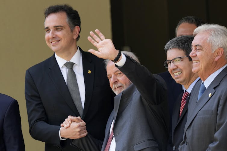 A short man in a suit salutes while surrounded by other men in suits, clasping a tall one's hand.