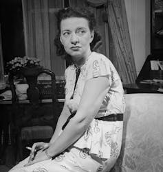 Retrato en blanco y negro de una mujer sentada, sosteniendo un cigarrillo y mirando lejos de la cámara.