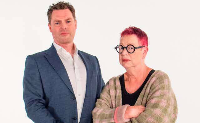 White background, man wearing suit on left, next to shorter woman on right with cropped red hair and black glasses