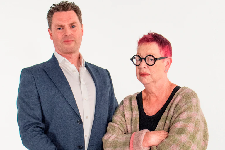 White background, man wearing suit on left, next to shorter woman on right with cropped red hair and black glasses