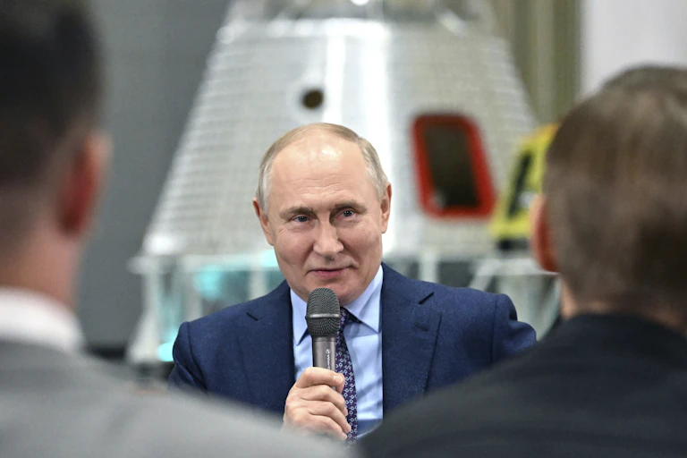 A man in a blue suit speaks into a microphone with a rocket behind him.