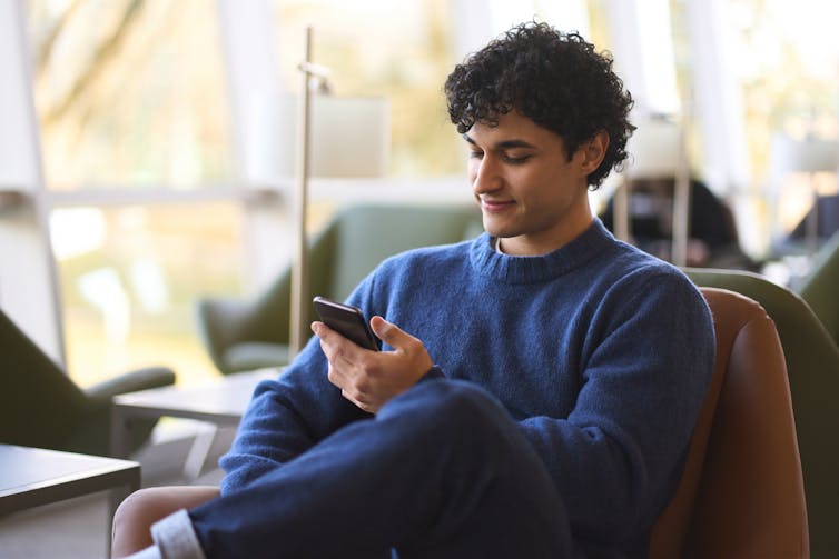 A young man with tan skin and dark curly hair sits in an armchair with one leg crossed over the other, casually scrolling on his mobile phone