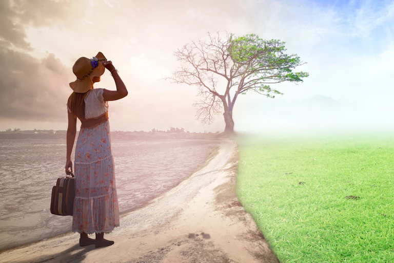 Woman in summer dress with hat and suitcase in foreground, path ahead and background spliut into left side grey barren field and right side green lush field with tree