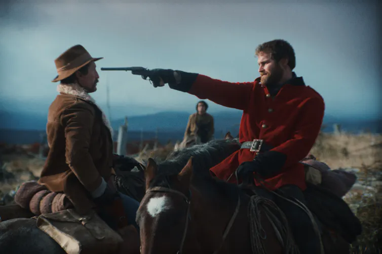 White man points a gun at the head of an indigenous man.
