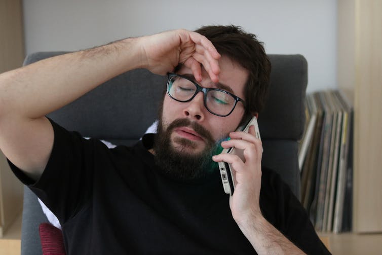 A man with dark hair and beard looking tired while listening to someone on the phone