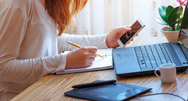 Une jeune femme assise à un bureau, devant un ordinateur portable, consulte son téléphone et écrit en même-temps dans un carnet.