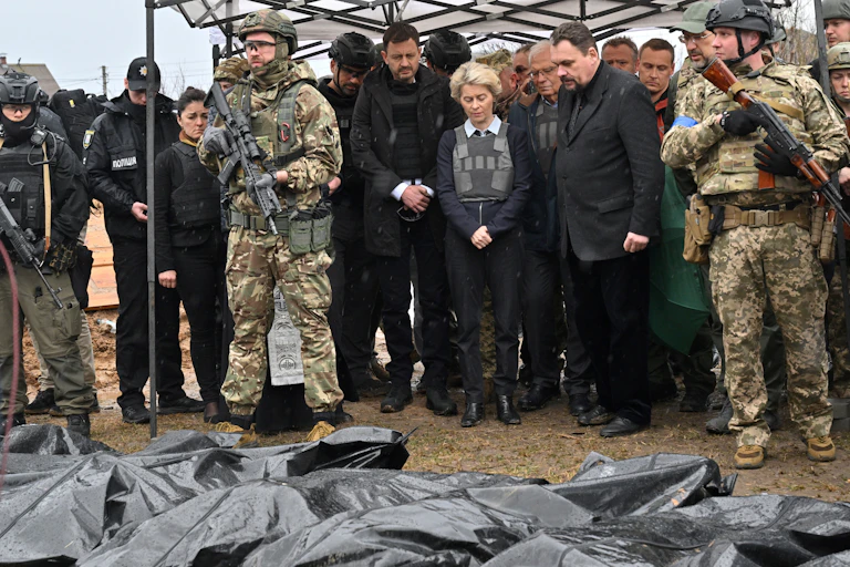 Plusieurs personnes dont Ursula von der Leyen se tiennent devant des corps placés dans des sacs noirs