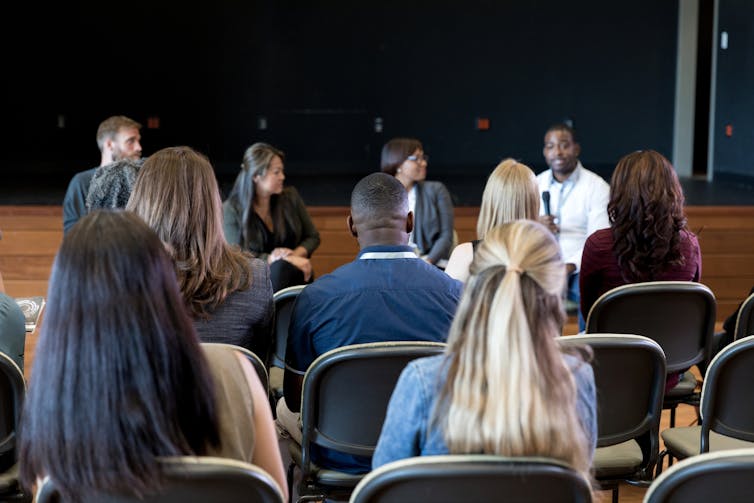 Two rows of seated people, seen from the back, listen to four people speaking as they face the audience.