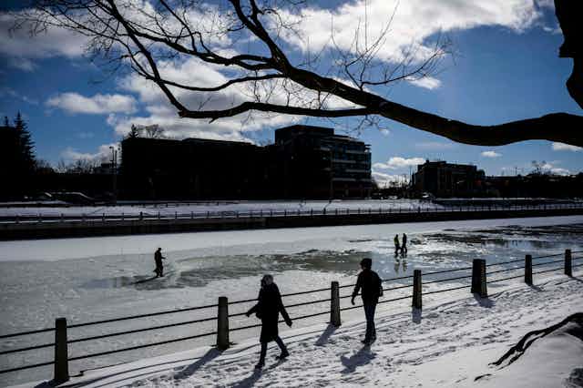 People walk past a partially frozen canal against a sunny sky.
