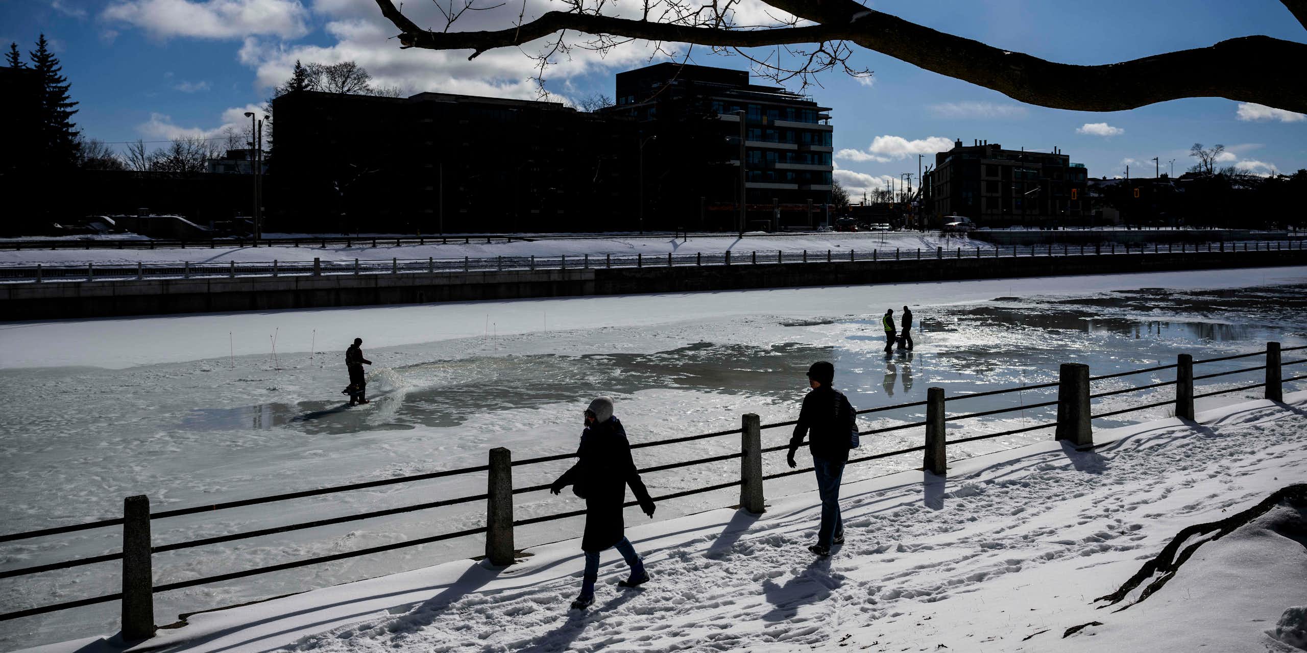 People walk past a partially frozen canal against a sunny sky.