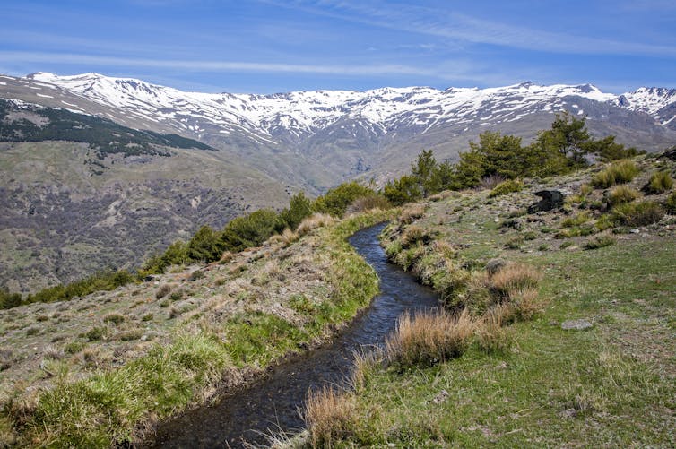 A canal running through a mountain side with snowy peaks in the background.