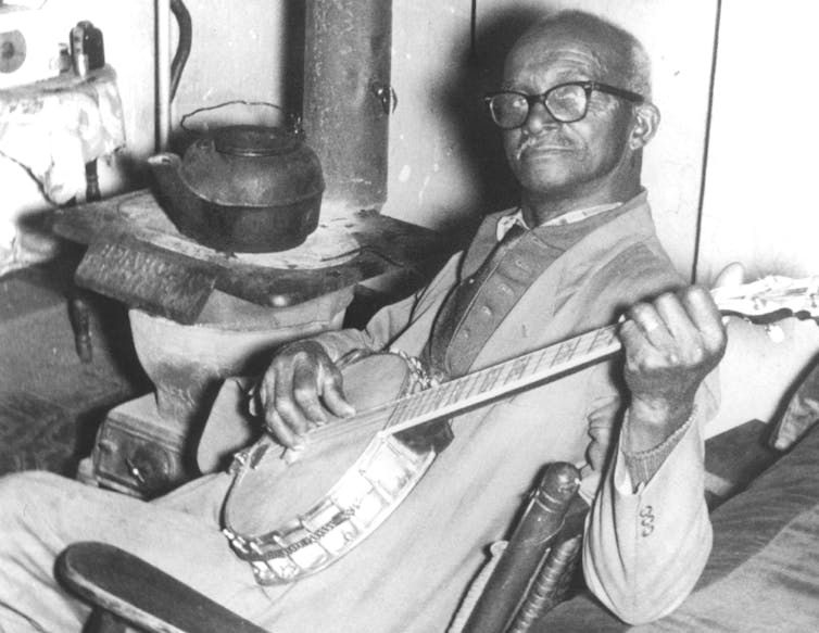 A black and white photograph of an older, balding Black man wearing glasses and sitting in a chair while strumming a banjo.