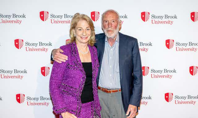 An older man in a jacket and no tie stands against a backdrop saying 'Stony Brook University' with his arm around a woman wearing a purple blazer and matching skirt