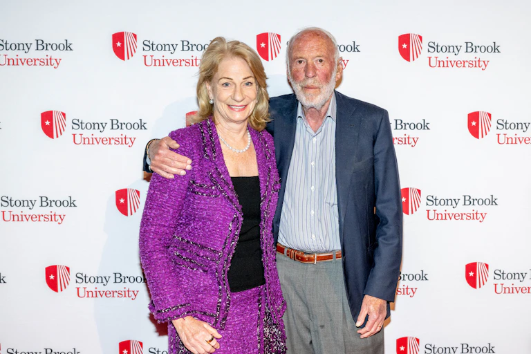 An older man in a jacket and no tie stands against a backdrop saying 'Stony Brook University' with his arm around a woman wearing a purple blazer and matching skirt