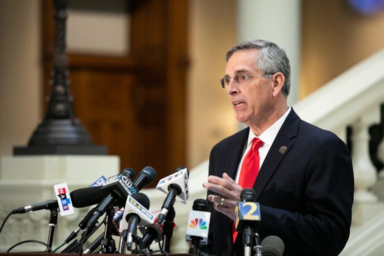 A man in a dark jacket with a red tie and white shirt talking behind many news organizations' microphones.