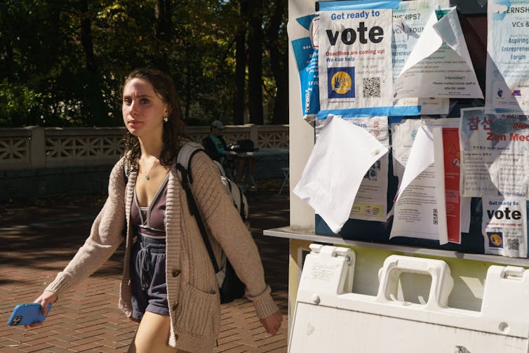 A young, white woman with brown hair wearing shorts and a beige cardigan walks past a bulletin board with flyers on it for vioting.