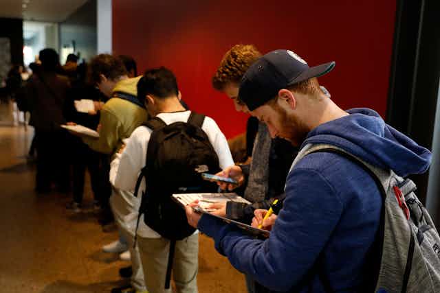 A row of young people stand inside a room with a red wall and look down at clipboards and white paper.